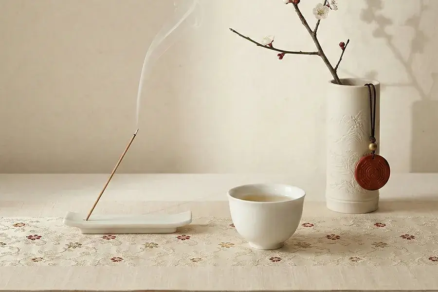 Minimalist Zen decor featuring burning traditional incense, an embroidered silk sachet, and a jade pendant against a textured beige wall.