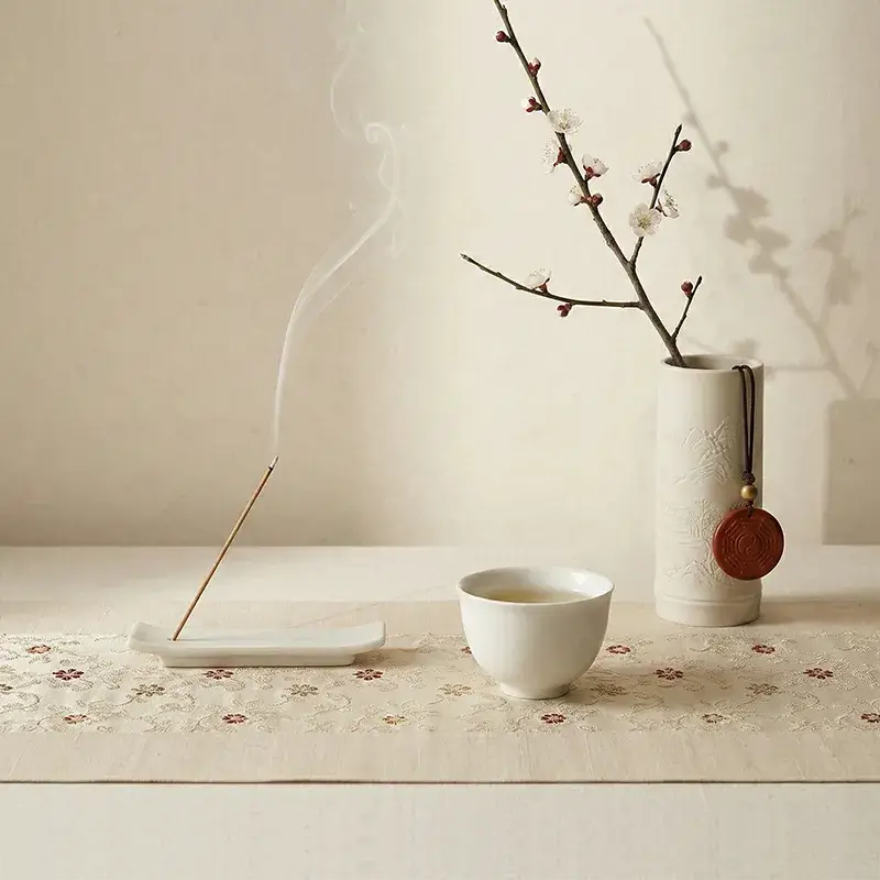 Minimalist Zen decor featuring burning traditional incense, an embroidered silk sachet, and a jade pendant against a textured beige wall.